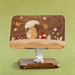 May include: A rectangular wooden tablet stand featuring an inlaid marquetry design of a large tan creature holding a leaf umbrella, a small red creature, and a white creature, surrounded by butterflies, flowers, and a mushroom on a dark wood base.