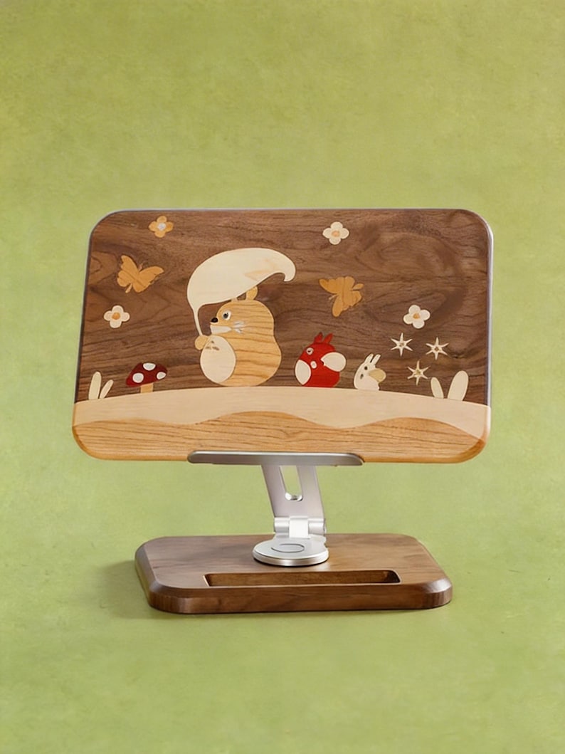 May include: A rectangular wooden tablet stand featuring an inlaid marquetry design of a large tan creature holding a leaf umbrella, a small red creature, and a white creature, surrounded by butterflies, flowers, and a mushroom on a dark wood base.