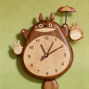 May include: A wooden wall clock shaped like a large brown creature with pointed ears, holding a small umbrella and two smaller light-coloured figures on its sides. The clock face is circular and light-toned with black numbers from 1 to 12.