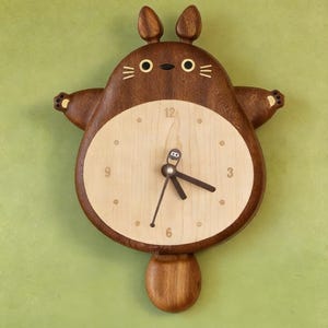 May include: A wooden wall clock shaped like a cartoon character, featuring a light wood clock face with black hands and a dark brown wooden frame. The clock has a whimsical design with ears and arms, and is mounted on a green wall.