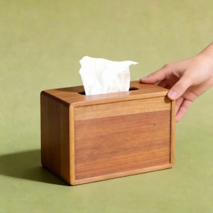 K&ouml;nnte beinhalten: Ein rechteckiger Taschentuchspender aus Holz in warmem Braunton. Die Box hat ein schlichtes, modernes Design mit abgerundeten Ecken und einer rechteckigen &Ouml;ffnung oben. Wei&szlig;e Taschent&uuml;cher sind sichtbar, die aus der &Ouml;ffnung ragen. Eine Hand h&auml;lt die Box.