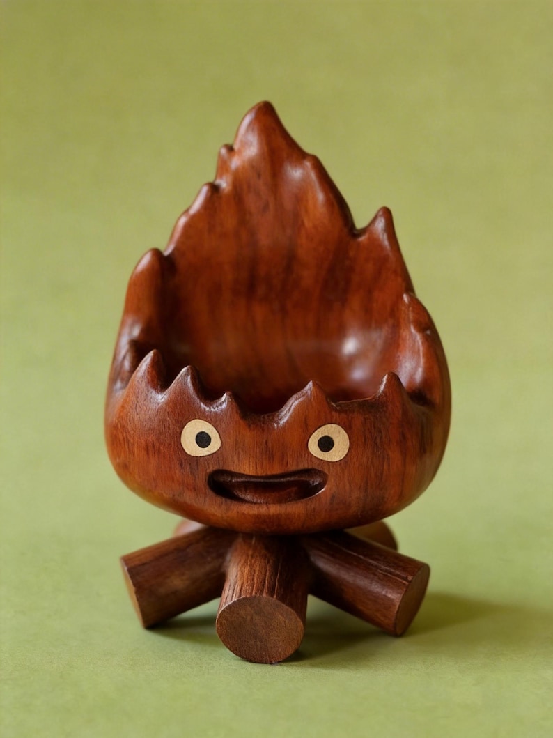 May include: A handcrafted wooden sculpture of a cartoon-like flame character. The sculpture is made of dark brown wood with a carved face featuring round eyes and a smiling mouth. The base has three wooden legs.