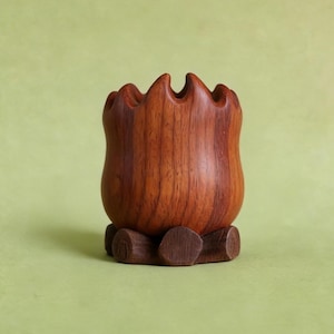 May include: A handcrafted wooden vase with a unique, wavy rim. The vase is a rich, warm brown color and sits on a small, round wooden base. The wood grain is visible, adding to the natural aesthetic. The vase is empty.