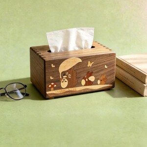May include: A rectangular wooden tissue box holder featuring an inlaid design of three cartoon creatures, one holding a leaf umbrella, alongside a mushroom and butterflies. The box is crafted from dark brown wood with lighter wood accents.
