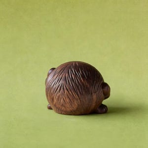Puede incluir: Peque&ntilde;a escultura redonda de madera de un animal, posiblemente un erizo o criatura similar, con detalles tallados. La escultura es marr&oacute;n y se asienta sobre un fondo verde. El tallado muestra la textura del pelaje del animal.