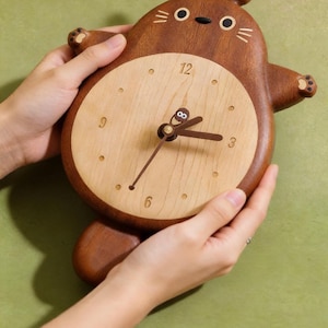 May include: A wooden wall clock shaped like a cartoon creature with a dark brown outer body and a light beige circular face. The clock features dark brown hands, a small decorative figure on the centre post, and numbers 3, 6, 9, and 12.