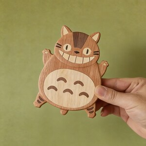 May include: Wooden coaster in the shape of a smiling cartoon cat with brown and light wood tones. The coaster has a large belly with curved markings and is held by a hand against a green background.