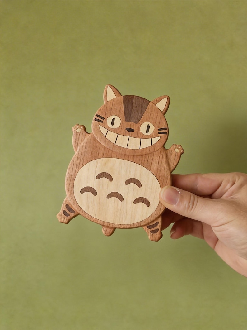 May include: Wooden coaster in the shape of a smiling cartoon cat with brown and light wood tones. The coaster has a large belly with curved markings and is held by a hand against a green background.