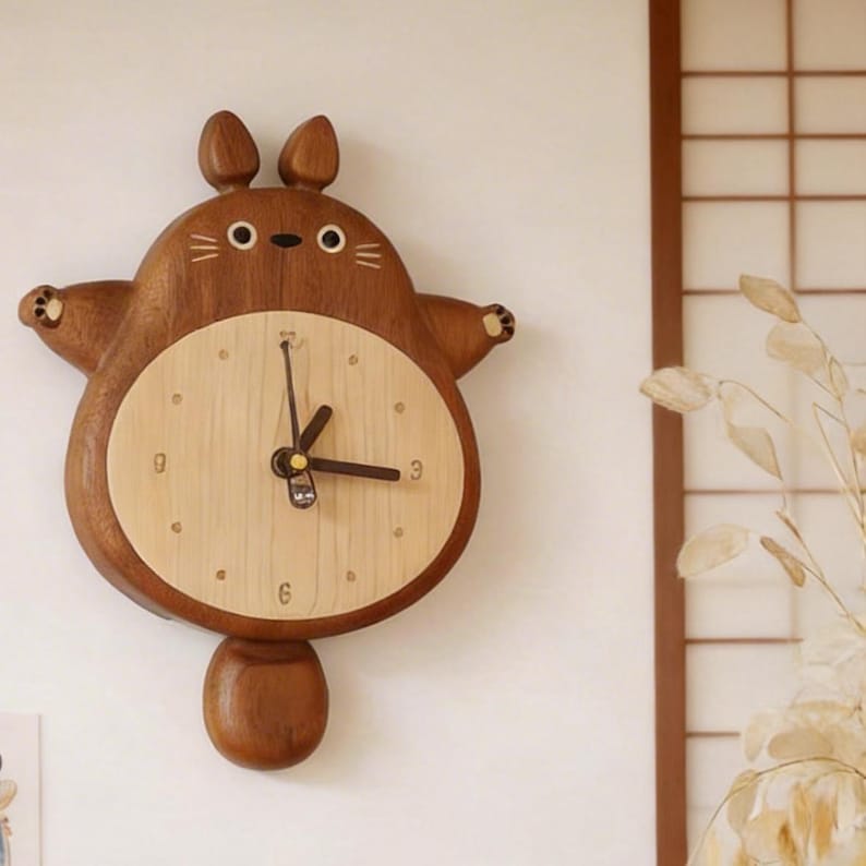 May include: A wooden wall clock shaped like a cartoon character with a round face and arms outstretched. The clock face is light-colored wood with black hands and numbers. The clock is made of dark brown wood and has a small pendulum.