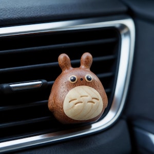 Puede incluir: Un clip de ventilación de coche de madera con forma de personaje de dibujos animados con cuerpo redondo, orejas pequeñas y cara sonriente. El clip es marrón con una cara de madera clara y está sujeto a una rejilla de ventilación del coche.