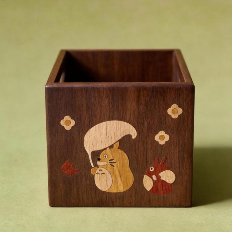May include: A square wooden box with a dark brown finish. The box is decorated with inlaid wood designs of cartoon characters, including a large, tan creature with a leaf-shaped umbrella and a smaller red and white creature. Small flower shapes are also inlaid.