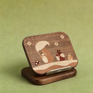May include: A rectangular wooden phone stand featuring an inlaid design of three cartoon creatures and a mushroom. The dark wood surface displays a light-colored inlay of a large creature holding a leaf, a smaller creature, and a tiny one, with flower accents.