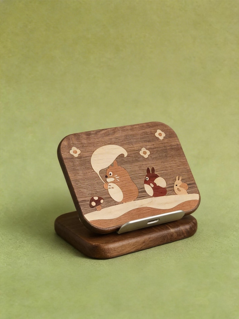May include: A rectangular wooden phone stand featuring an inlaid design of three cartoon creatures and a mushroom. The dark wood surface displays a light-colored inlay of a large creature holding a leaf, a smaller creature, and a tiny one, with flower accents.