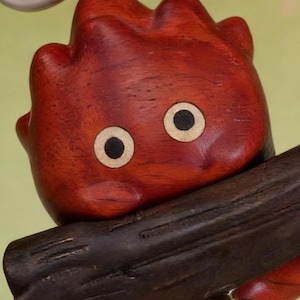 May include: A wooden sculpture of a red, cartoon-like creature with large, round eyes. The figure has a textured surface and is perched on a dark brown wooden beam. The eyes are beige with black pupils, and the creature's body is a deep reddish-brown.