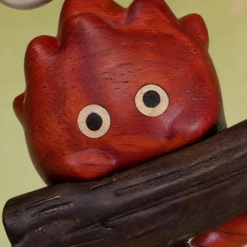 May include: A wooden sculpture of a red, cartoon-like creature with large, round eyes. The figure has a textured surface and is perched on a dark brown wooden beam. The eyes are beige with black pupils, and the creature's body is a deep reddish-brown.