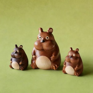 May include: Three wooden figurines of a bear-like creature with rounded bodies, small ears, and light-colored bellies. The figures are in varying sizes, with the largest in the center and the smallest on the left. The background is a solid green.