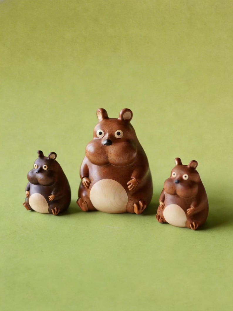 May include: Three wooden figurines of a bear-like creature with rounded bodies, small ears, and light-colored bellies. The figures are in varying sizes, with the largest in the center and the smallest on the left. The background is a solid green.
