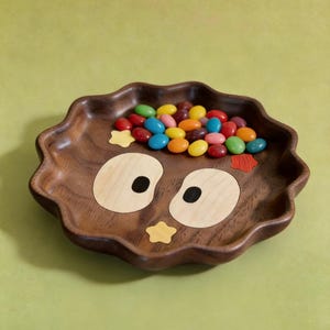 May include: A wooden owl-shaped tray with a wavy edge, filled with colourful sweets. The tray is made of dark brown wood, with light wood and black accents for the owl's eyes and a yellow star. A fun and decorative piece.