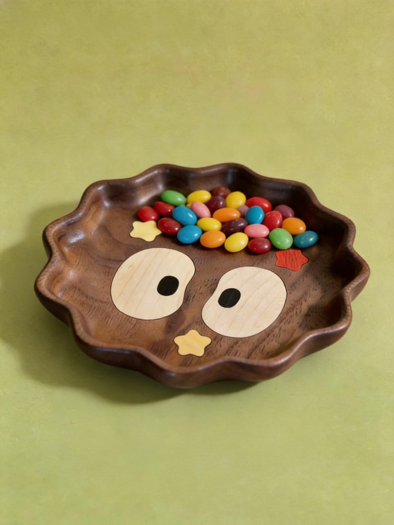 May include: A wooden owl-shaped tray with a wavy edge, filled with colourful sweets. The tray is made of dark brown wood, with light wood and black accents for the owl's eyes and a yellow star. A fun and decorative piece.