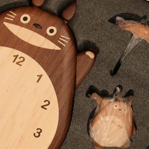 May include: A wooden wall clock shaped like a cartoon creature with a dark brown body and a light beige face featuring black numerals from 1 to 12. The clock is packaged in grey foam alongside a small umbrella accessory and a matching figurine.
