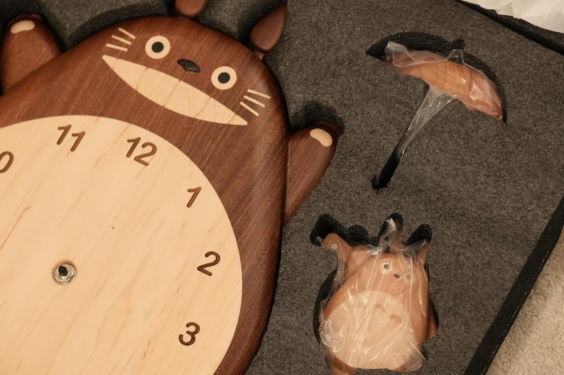 May include: A wooden wall clock shaped like a cartoon creature with a dark brown body and a light beige face featuring black numerals from 1 to 12. The clock is packaged in grey foam alongside a small umbrella accessory and a matching figurine.