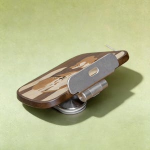 May include: A wooden phone stand featuring a dark and light wood inlay design of a dog's face. The stand includes a silver-colored metal adjustable hinge and a circular base, allowing for multiple viewing angles for a mobile device.