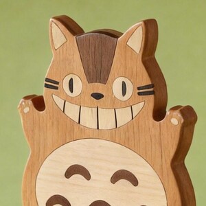May include: A wooden sculpture of a smiling cat-like creature with large eyes and a wide grin. The sculpture is made of various shades of wood, with a light brown body, darker brown accents, and a lighter belly.