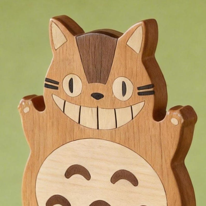May include: A wooden sculpture of a smiling cat-like creature with large eyes and a wide grin. The sculpture is made of various shades of wood, with a light brown body, darker brown accents, and a lighter belly.