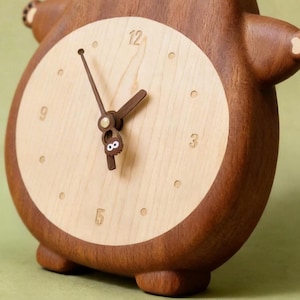 May include: A wooden clock shaped like a bear with a light-coloured circular face. The clock features dark brown hour and minute hands, with a small brown bear-shaped detail on the minute hand. The numbers 12, 3, 6, and 9 are engraved.