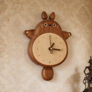 May include: A wooden wall clock shaped like a cartoon creature with two ears, a round body, and a tail. The clock face is a lighter wood tone with simple hour markers and three dark hands. The outer frame is a darker brown wood with carved details.
