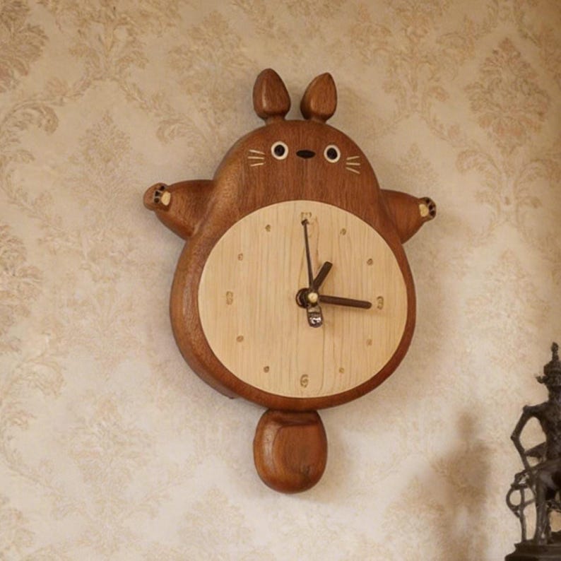 May include: A wooden wall clock shaped like a cartoon creature with two ears, a round body, and a tail. The clock face is a lighter wood tone with simple hour markers and three dark hands. The outer frame is a darker brown wood with carved details.