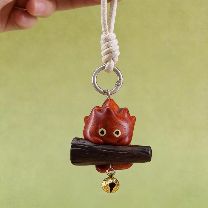 May include: A wooden keychain featuring a stylized, red, flame-like character with two small, round eyes. The character is perched on a dark brown wooden log, with a small gold-colored bell hanging below. The keychain has a cream-colored rope and a silver-colored ring.