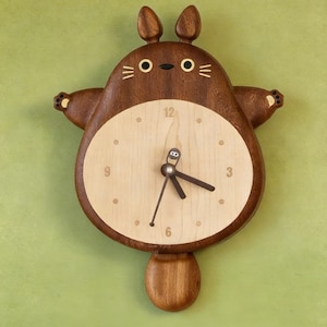 May include: A wooden wall clock shaped like a cartoon creature with two ears, a round light-coloured face, and a dark brown body. The clock features a central dial with numbers 3, 6, 9, and 12, dark brown hands, and a small pendulum shaped like a tail.