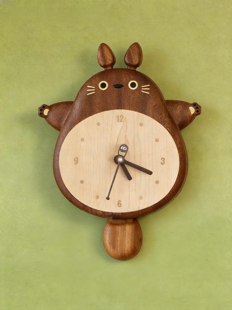 May include: A wooden wall clock shaped like a cartoon creature with two ears, a round light-coloured face, and a dark brown body. The clock features a central dial with numbers 3, 6, 9, and 12, dark brown hands, and a small pendulum shaped like a tail.