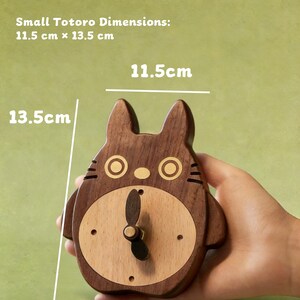 May include: A wooden Totoro-shaped clock, 11.5 cm x 13.5 cm, with a light wood face and dark wood body. The clock has a black clock hand and is designed to be wall-mounted or displayed on a tabletop. The clock has a light green background.