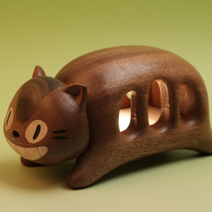 May include: A wooden cat-shaped figurine with a smiling face and three oval openings. The figurine is made of dark brown wood with lighter wood accents for the eyes and teeth. The cat has small ears and is positioned on a light green surface.