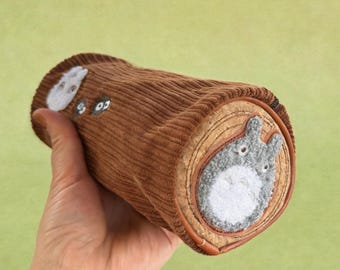 May include: A brown corduroy pencil case shaped like a log, featuring embroidered Totoro characters. The ends of the case showcase a cross-section design with a gray Totoro. The case is held in a hand against a light green background.