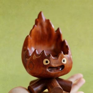 May include: A handcrafted wooden decorative bowl in the shape of a cartoon character. The bowl is made of dark brown wood, with a carved flame-like top and a smiling face. The bowl is supported by three wooden legs.