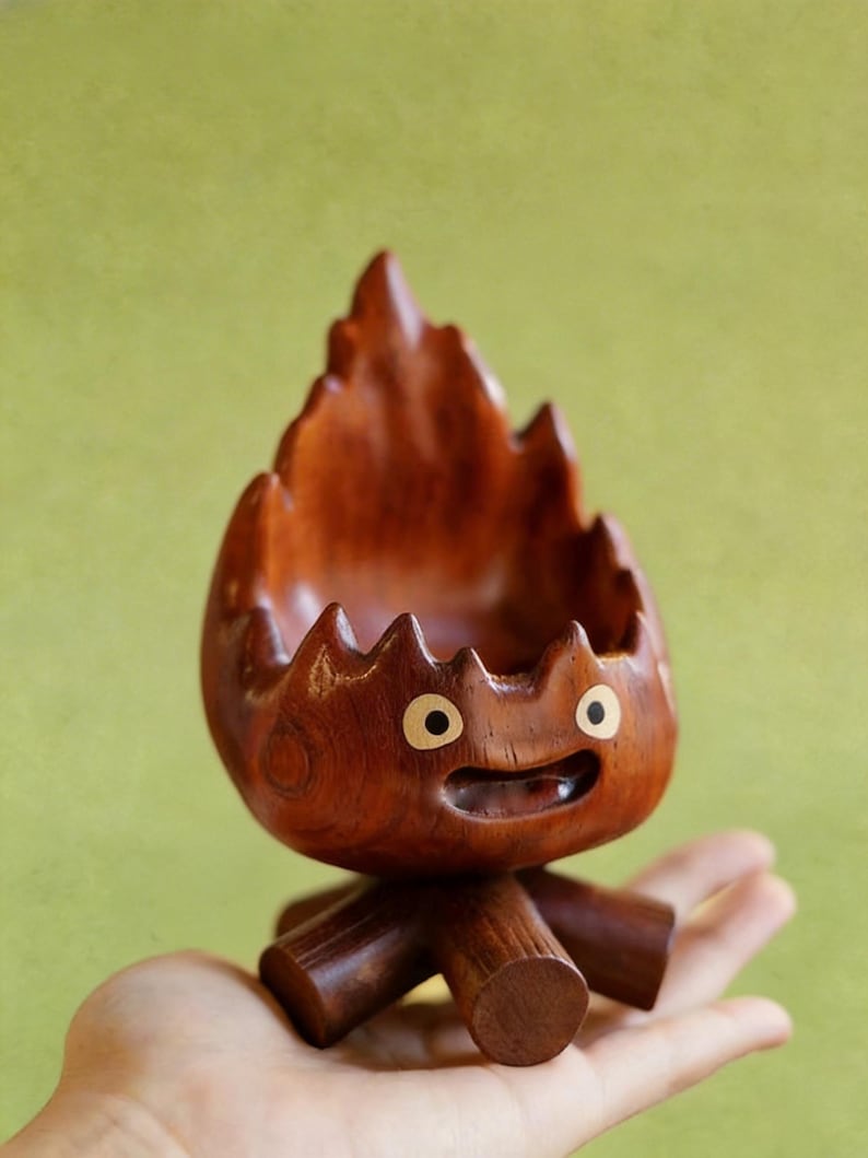 May include: A handcrafted wooden decorative bowl in the shape of a cartoon character. The bowl is made of dark brown wood, with a carved flame-like top and a smiling face. The bowl is supported by three wooden legs.