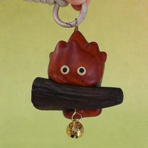 May include: A wooden keychain featuring a stylized, flame-shaped character with two white eyes and a dark brown log. A small gold-colored bell hangs below. The keychain is attached to a white cord and a silver-colored ring.