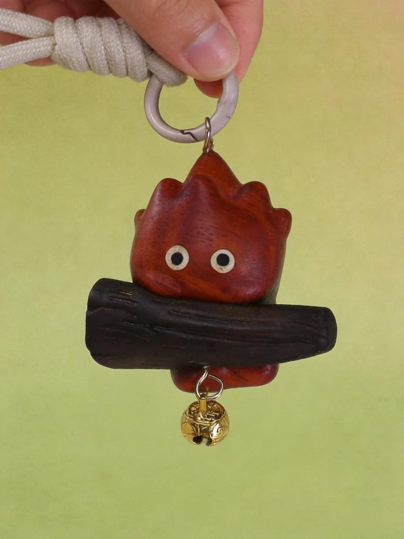 May include: A wooden keychain featuring a stylized, flame-shaped character with two white eyes and a dark brown log. A small gold-colored bell hangs below. The keychain is attached to a white cord and a silver-colored ring.