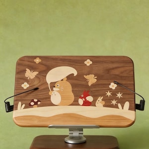 May include: A rectangular wooden book stand featuring an inlaid marquetry design of a cartoon creature holding a leaf umbrella, accompanied by a smaller red creature, a rabbit, butterflies, flowers, and a mushroom on a dark wood grain surface.
