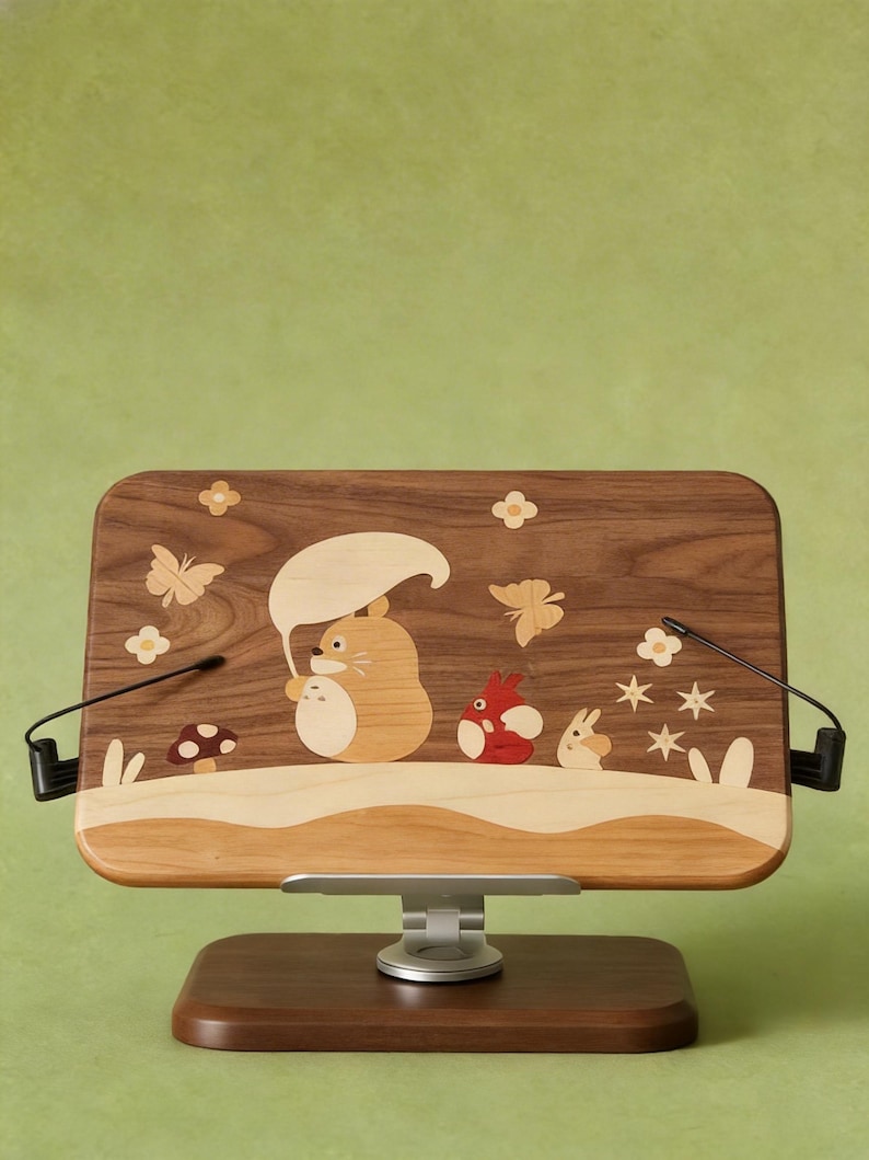 May include: A rectangular wooden book stand featuring an inlaid marquetry design of a cartoon creature holding a leaf umbrella, accompanied by a smaller red creature, a rabbit, butterflies, flowers, and a mushroom on a dark wood grain surface.