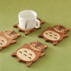 May include: Wooden coasters in the shape of a smiling cat, with a white marble mug on one. The coasters are brown with lighter wood accents, and the mug has a black marble pattern. The coasters are on a green surface.