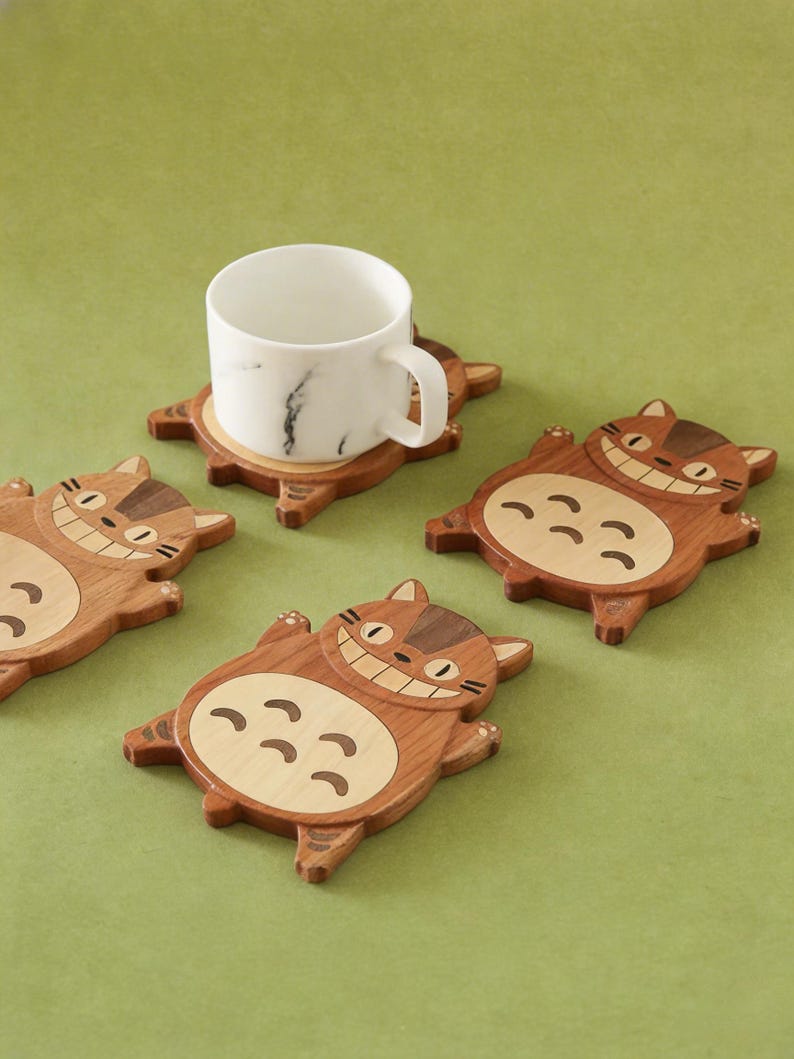May include: Wooden coasters in the shape of a smiling cat, with a white marble mug on one. The coasters are brown with lighter wood accents, and the mug has a black marble pattern. The coasters are on a green surface.