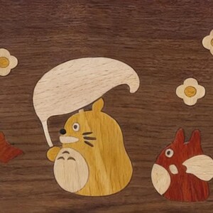 May include: Wooden inlay art featuring a cartoon character holding a leaf umbrella, with a smaller character to the right. The artwork includes flower and leaf accents, all set against a dark wood background. The characters are in shades of yellow and red.