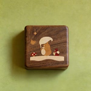 May include: Square wooden box with a detailed forest scene. The box features a squirrel under a mushroom, with other forest elements inlaid in various wood tones. The box is made of dark brown wood.