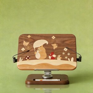 May include: A rectangular wooden book stand featuring an inlaid design of a cartoon creature holding a leaf umbrella, accompanied by a smaller red creature, a rabbit, butterflies, flowers, and a mushroom. The stand has a metal base and two arms.