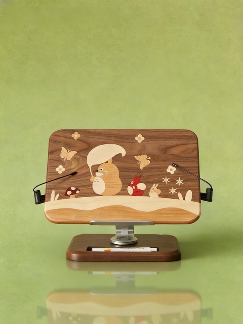 May include: A rectangular wooden book stand featuring an inlaid design of a cartoon creature holding a leaf umbrella, accompanied by a smaller red creature, a rabbit, butterflies, flowers, and a mushroom. The stand has a metal base and two arms.