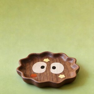 May include: A decorative wooden tray with a wavy edge. The tray is brown with inlaid details forming a cartoon face: large white eyes with black pupils, an orange beak, and yellow star accents. The tray is set against a solid green background.